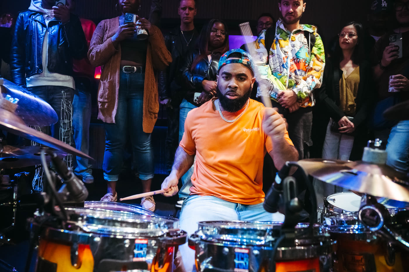 A drummer mid-strike with the crowd standing in a full circle around the kit in the Live Room.